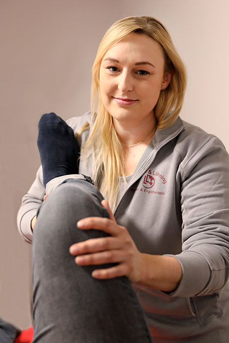 Eine Frau hält das angewinkelte Bein eines Patienten und lächelt dabei in die Kamera.