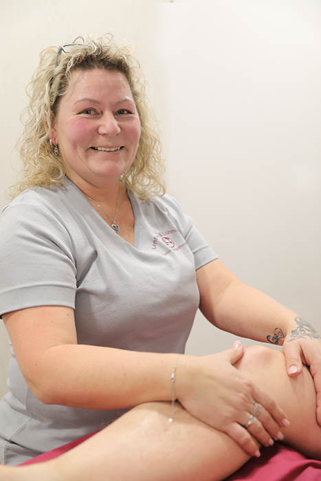 Eine Frau legt ihre Hände auf das Knie einer Patientin und lächelt dabei in die Kamera.