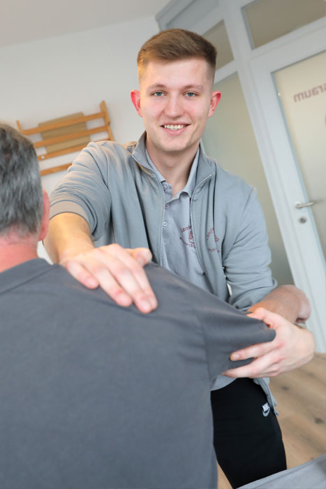 Junger Mann behandelt die Schulter eines Patienten mit Blick in die Kamera.