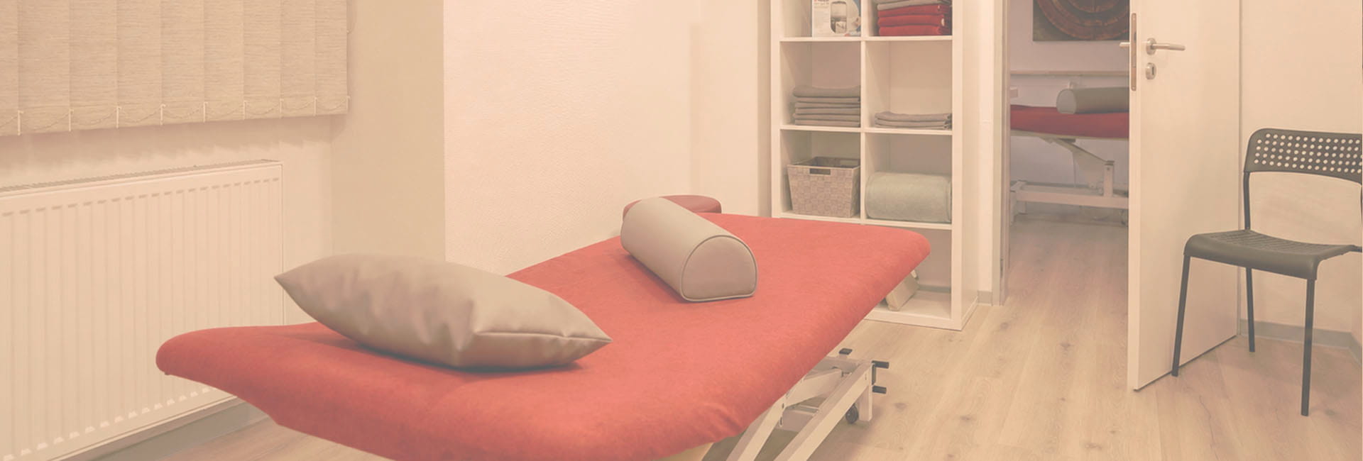 Behandlungsraum mit roter Therapieliege, Kissen, Regal und Sitzgelegenheit